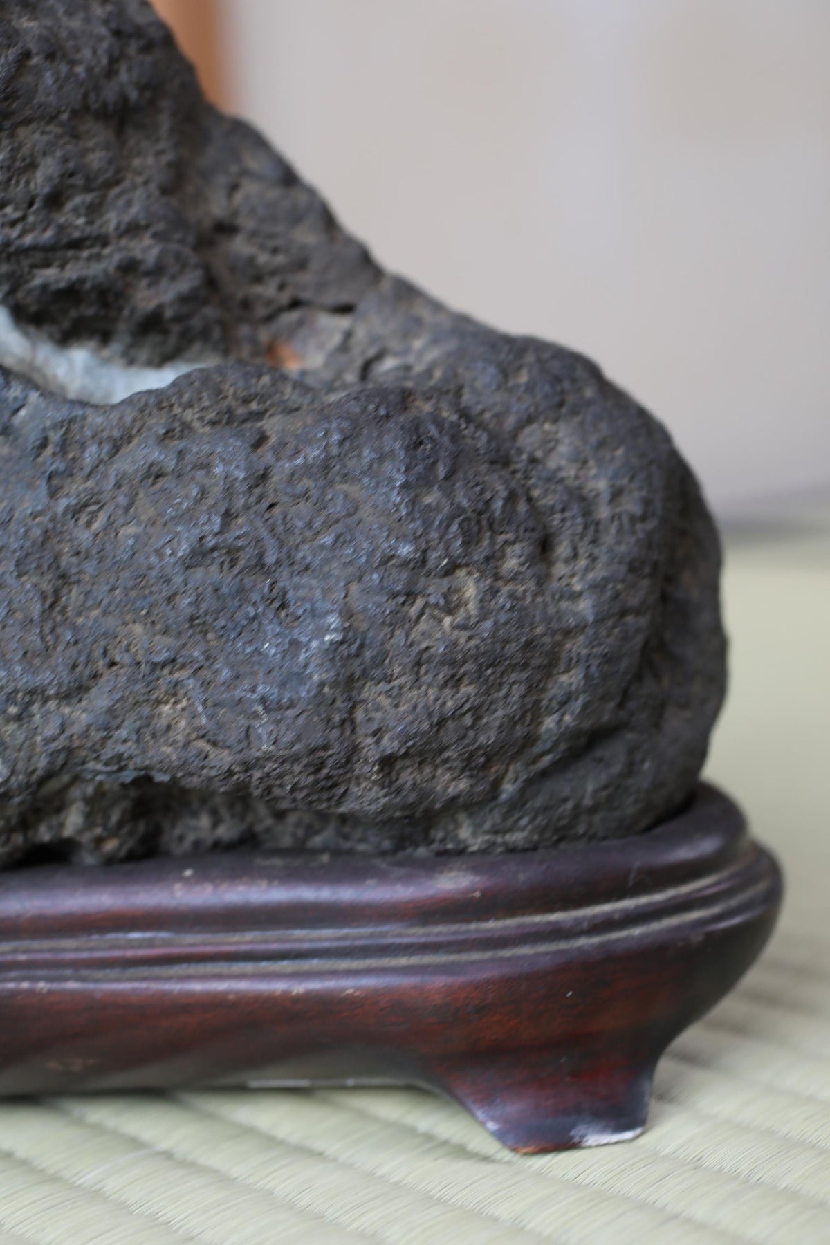 A close-up image of a dark, textured rock or stone, possibly a piece of art or sculpture, placed on a wooden base or stand.