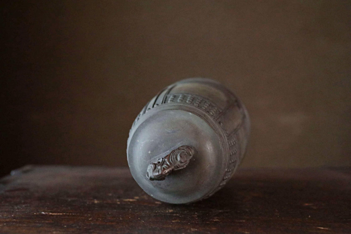 A gray metal object with a decorative design on the top sits on a wooden surface against a dark background.