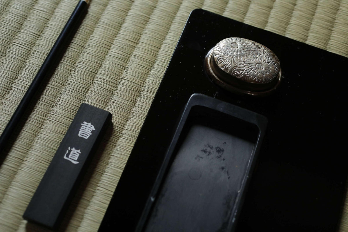 a close-up view of a black object with a silver flower design on it, placed on a woven mat. There is also a black object with white text on it, and a black object with a silver flower design on it.