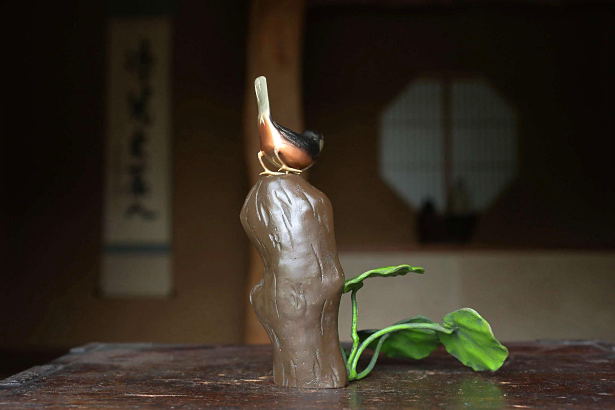 A ceramic bird figurine perched on a tree stump, with a green leaf attached to it, sits on a wooden surface.