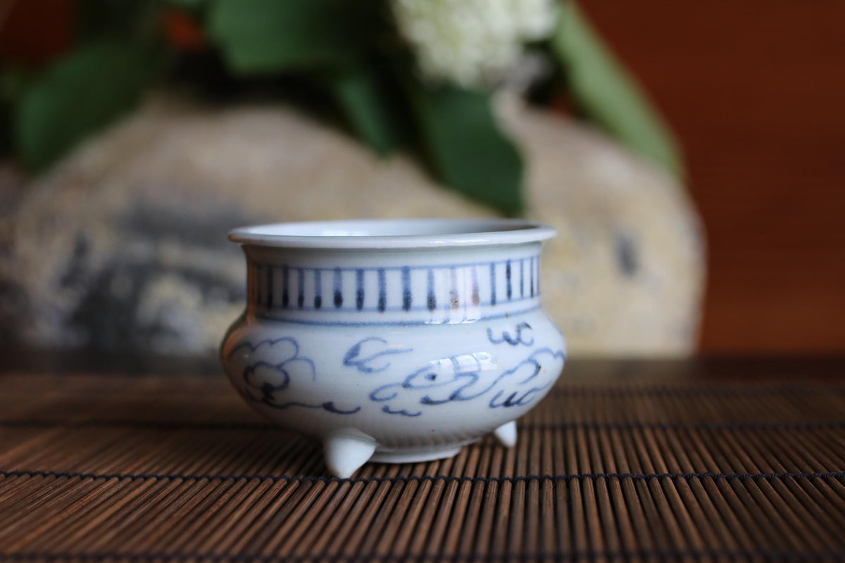 A white ceramic bowl with blue and black designs sits on a bamboo mat, with a blurred background of greenery.