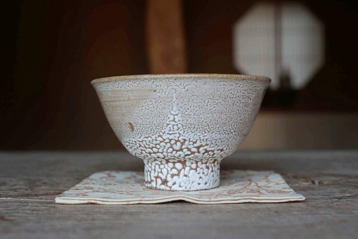 A white ceramic bowl with a speckled pattern sits on a white cloth napkin on a wooden surface.