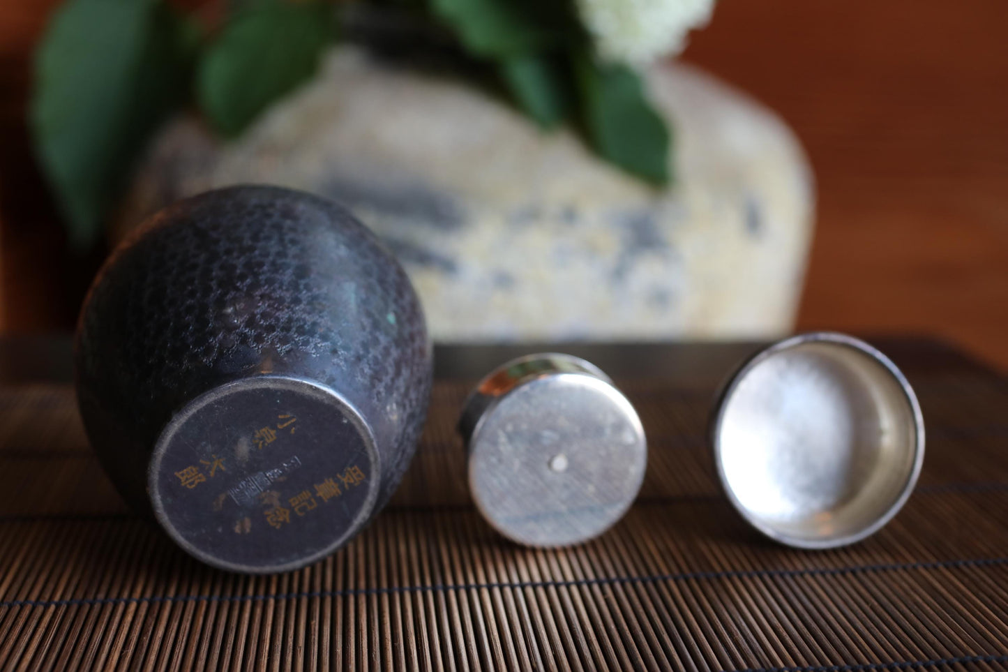 three metal objects, including a small metal container and two metal containers, placed on a wooden surface. In the background, there is a vase with green leaves.