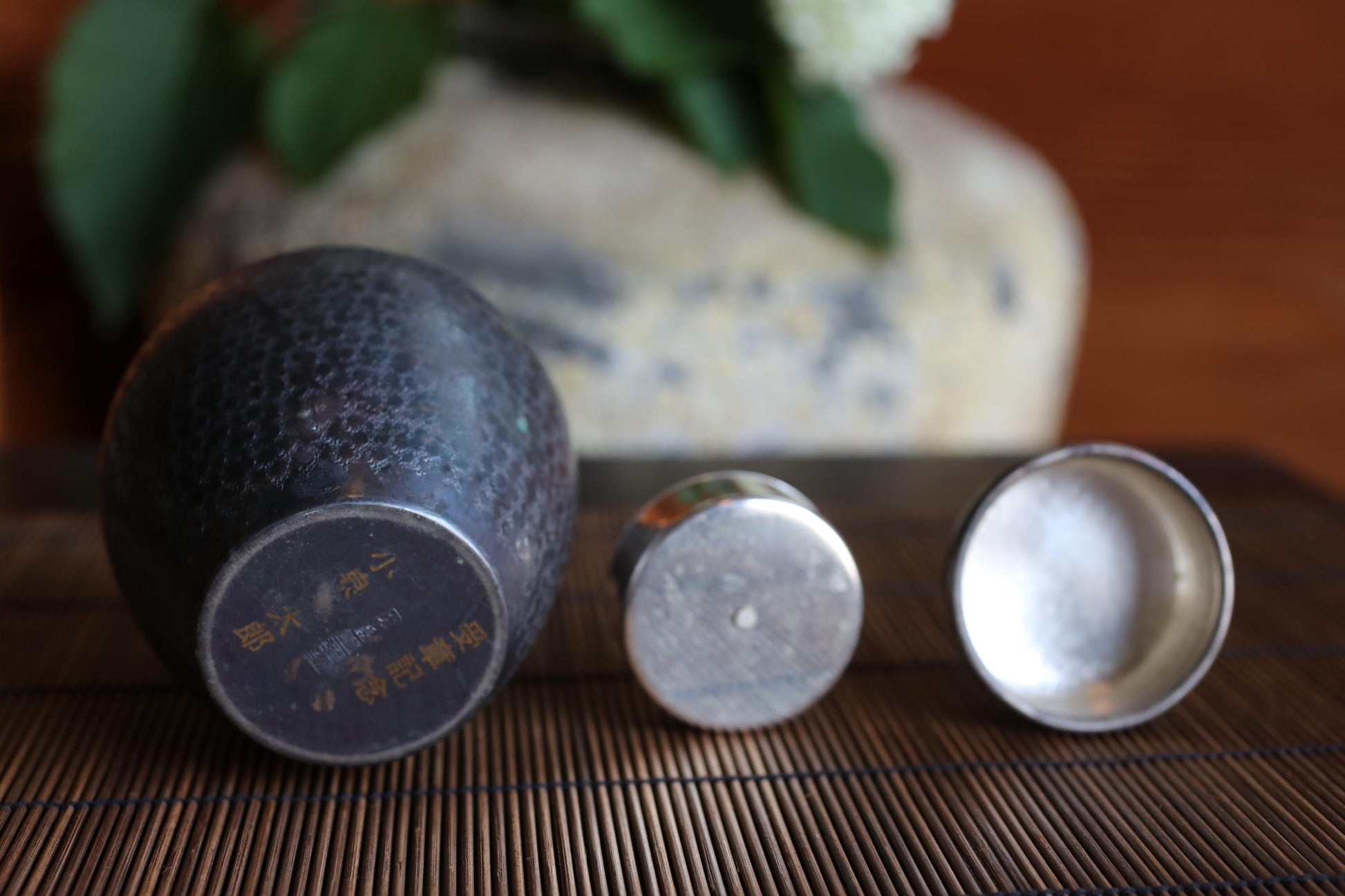 three metal objects, including a small metal container and two metal containers, placed on a wooden surface. In the background, there is a vase with green leaves.