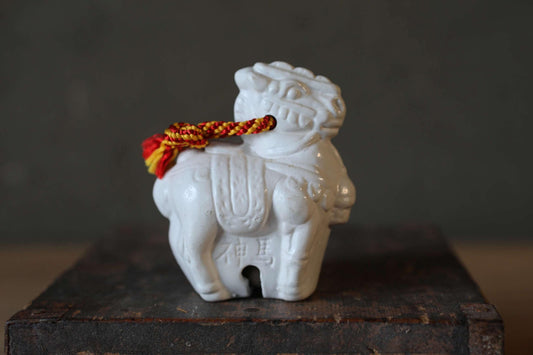 A white ceramic figurine of a lion with a red and yellow tassel is displayed on a wooden surface.