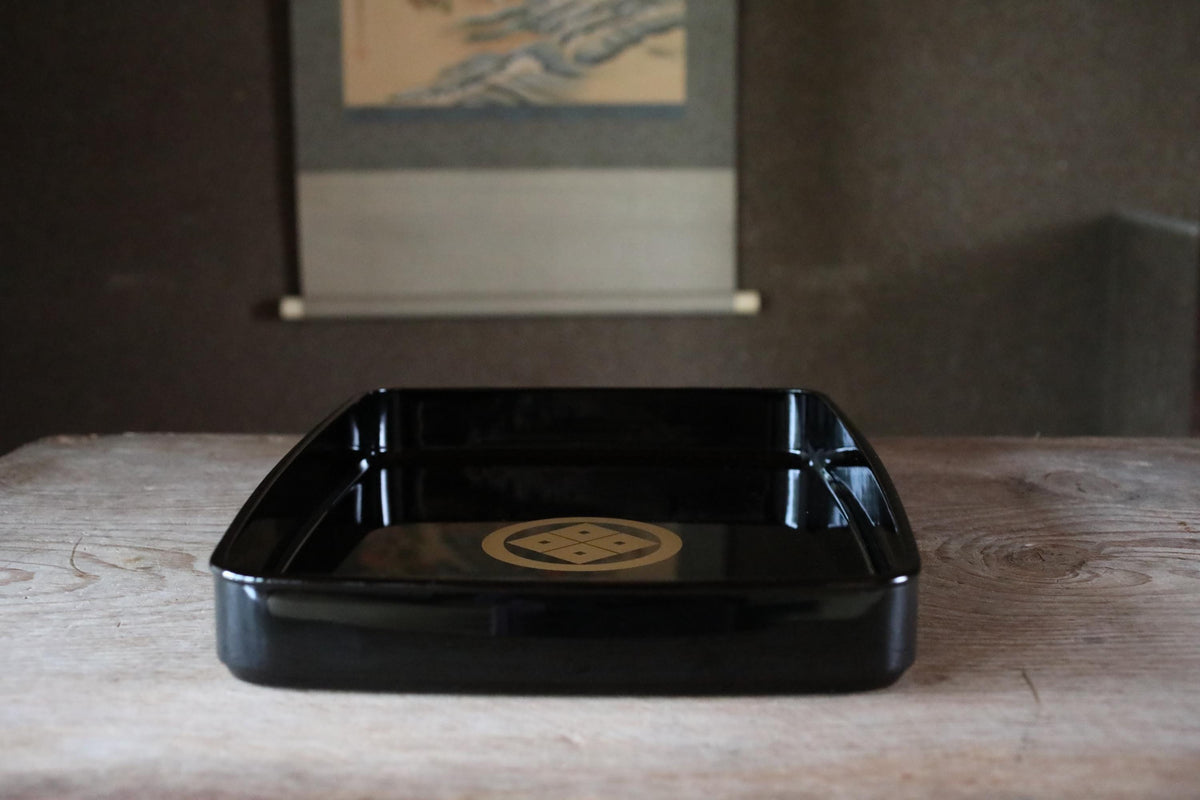 A black rectangular tray with a gold emblem on it, placed on a wooden surface.