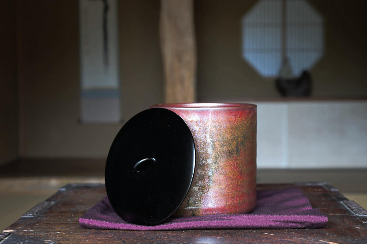 A red and black ceramic jar with a black lid sits on a purple cloth on a wooden surface, with a blurred background suggesting an indoor setting.