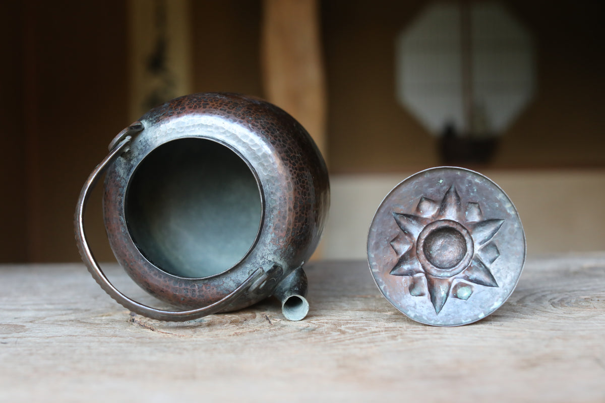 A copper-colored kettle with a star-shaped lid sits on a wooden surface, with a blurred background suggesting an indoor setting.