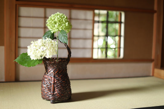 A small woven basket containing white hydrangeas and a green flower sits on a wooden surface, with a window and wooden wall in the background.