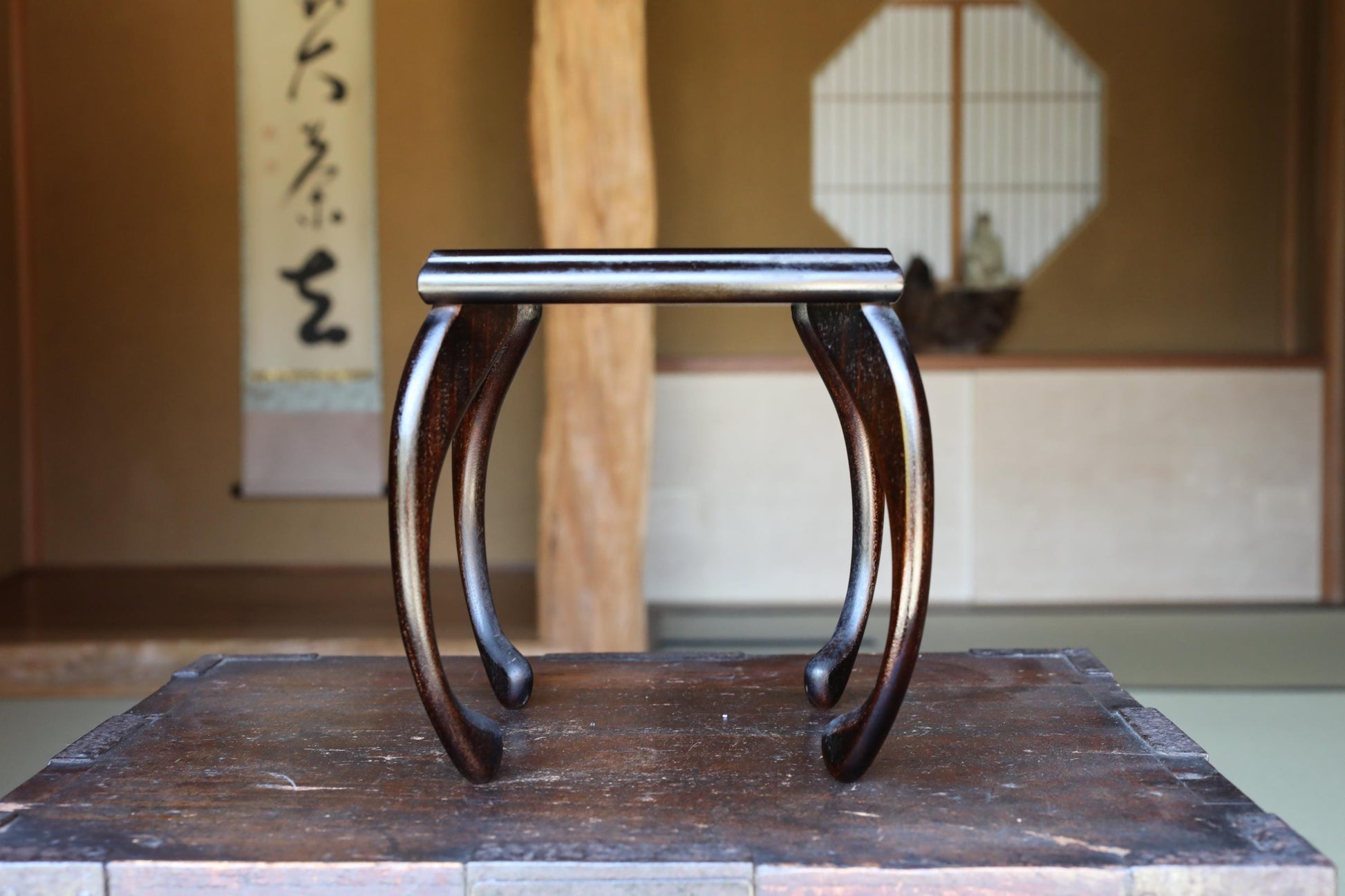 A wooden table with a curved top and legs, placed in a room with a traditional Japanese-style wall hanging and a window.