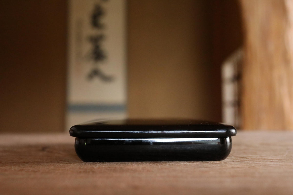 A black smartphone rests on a wooden surface, with a blurred background that includes a wall with Japanese characters.