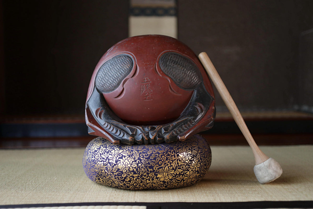 A red ceramic bowl with intricate designs sits on a blue and gold patterned base, accompanied by a wooden drumstick.