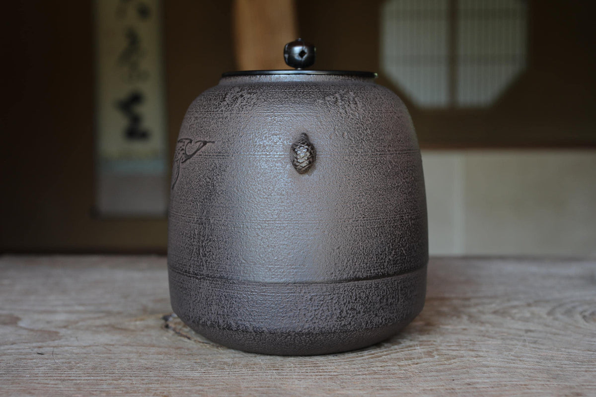 A ceramic teapot with a black lid sits on a wooden surface, with a blurred background that includes a window and a wall.