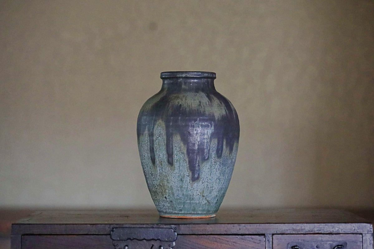 A dark blue ceramic vase with a textured surface sits on a wooden surface against a plain background.