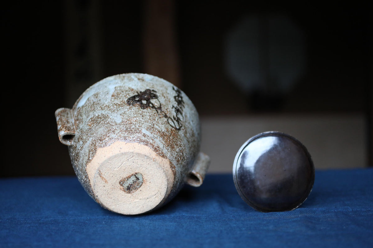 An old, weathered ceramic jar with a distinctive design sits on a blue surface, accompanied by a round, reflective lid.