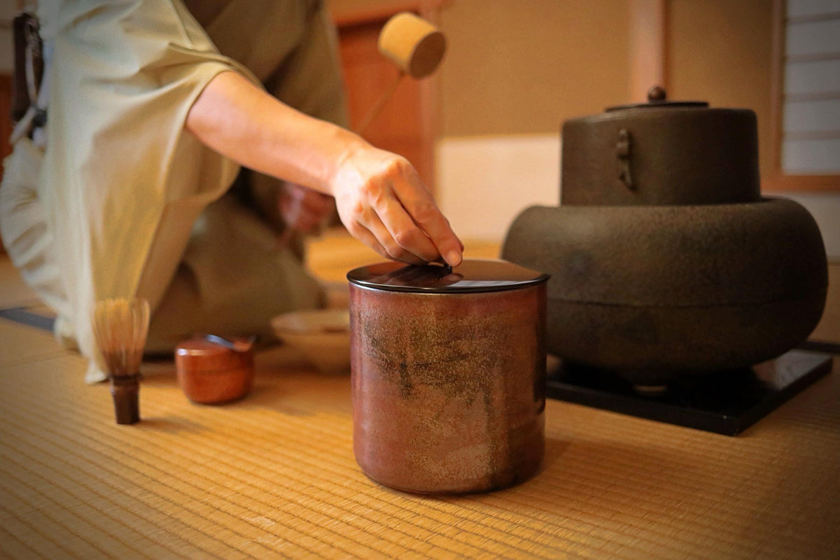 A person is carefully placing a small object into a red ceramic container on a wooden surface.