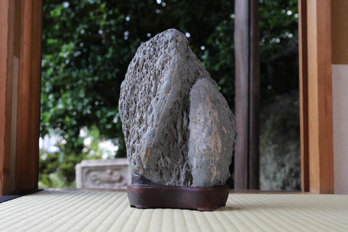 A large, gray rock sits on a wooden base in a serene garden setting, surrounded by lush greenery.