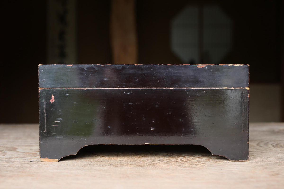 A black wooden box with a weathered appearance sits on a wooden surface.