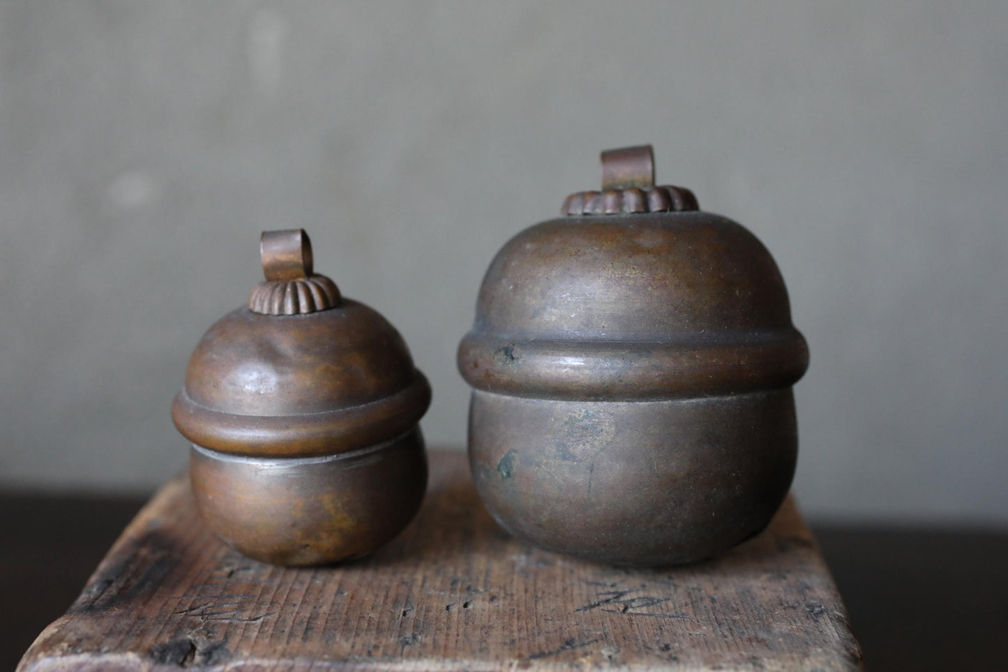 Two antique brass containers with round tops sit on a wooden surface against a gray background.