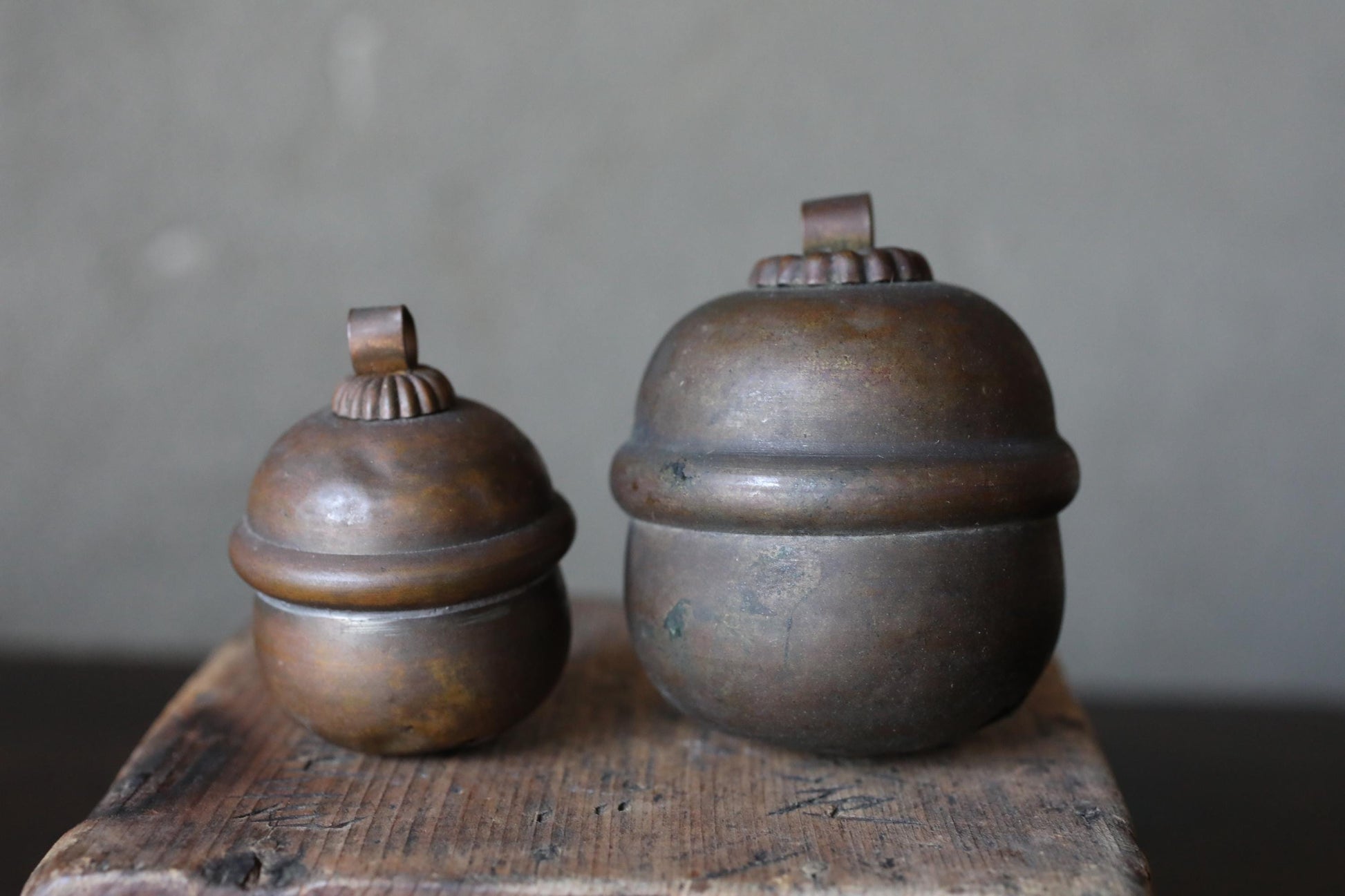 Two antique brass containers with round tops sit on a wooden surface against a gray background.