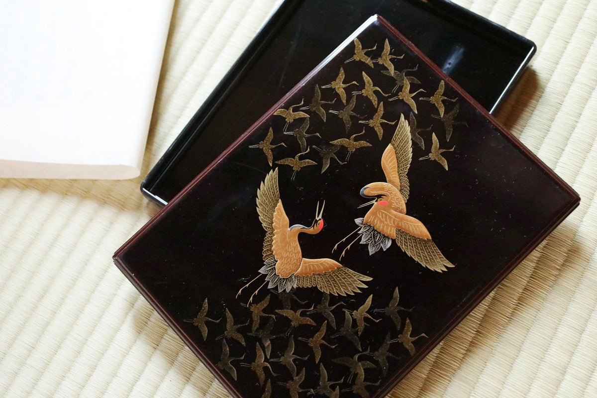 a black tray with a decorative design of two cranes in flight, surrounded by a pattern of small birds.