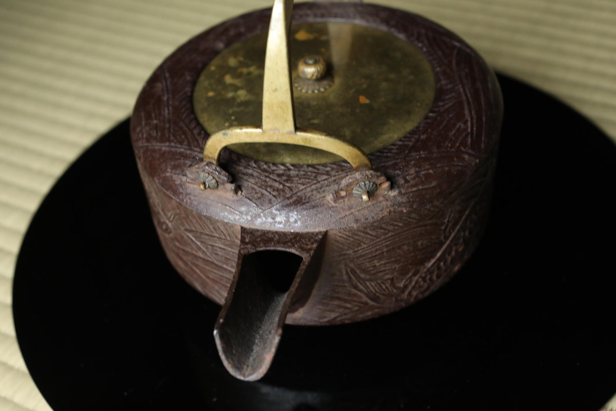 A close-up view of a dark brown, intricately designed teapot with a gold-colored lid and handle, resting on a black surface.