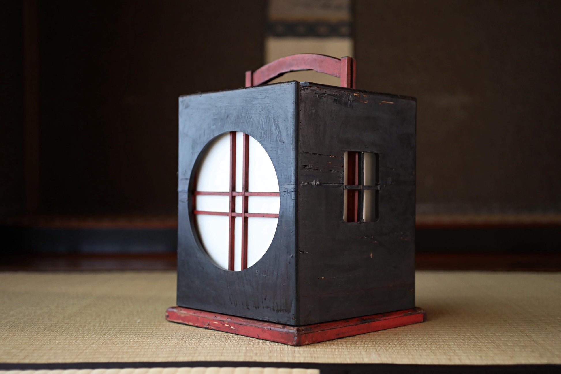 A black lantern with a red handle, featuring a circular cutout in the center, is displayed on a beige surface.