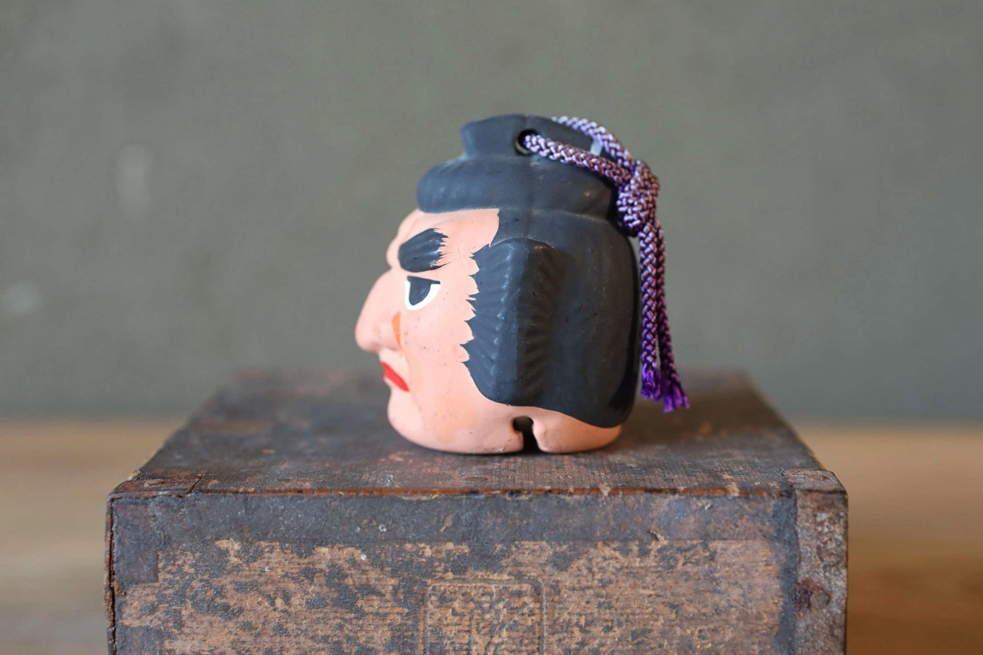 A ceramic figurine of a person's head with a black hat and purple tassel is displayed on a wooden crate.
