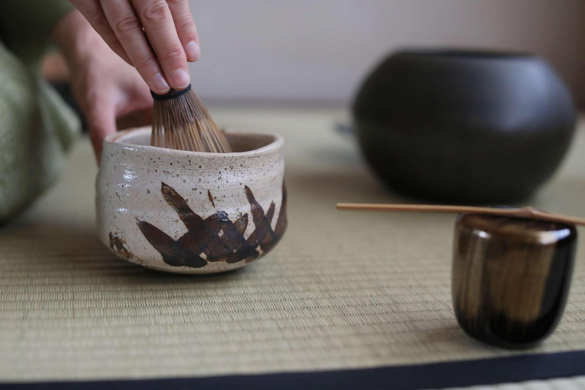 A hand is using a whisk to stir something in a ceramic bowl, which is placed on a woven mat. In the background, there are two other ceramic bowls, one larger and one smaller, as well as a wooden spoon.