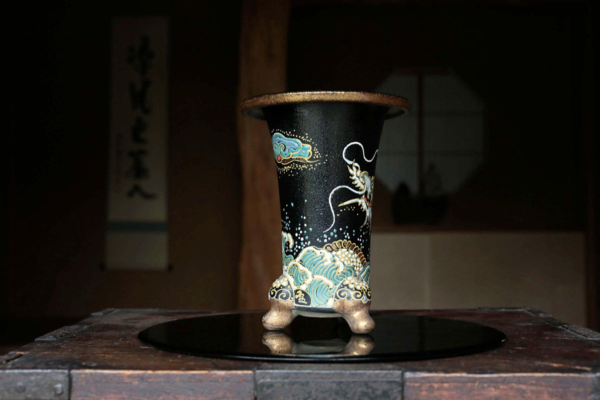 A black vase with intricate blue and gold designs sits on a wooden surface, with a dark background that includes a window and a sign with Japanese characters.