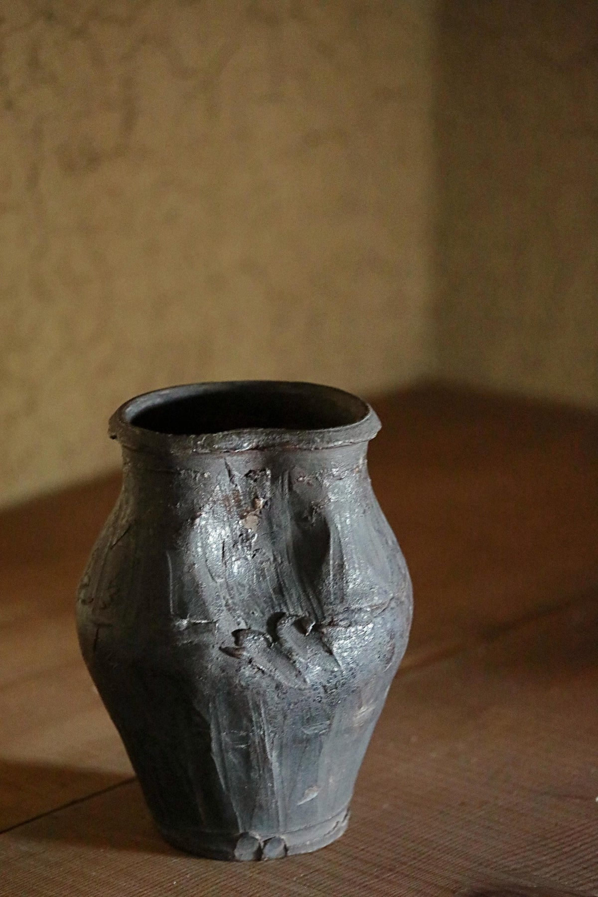 An old, weathered vase with a dark, textured surface sits on a wooden surface against a plain wall.