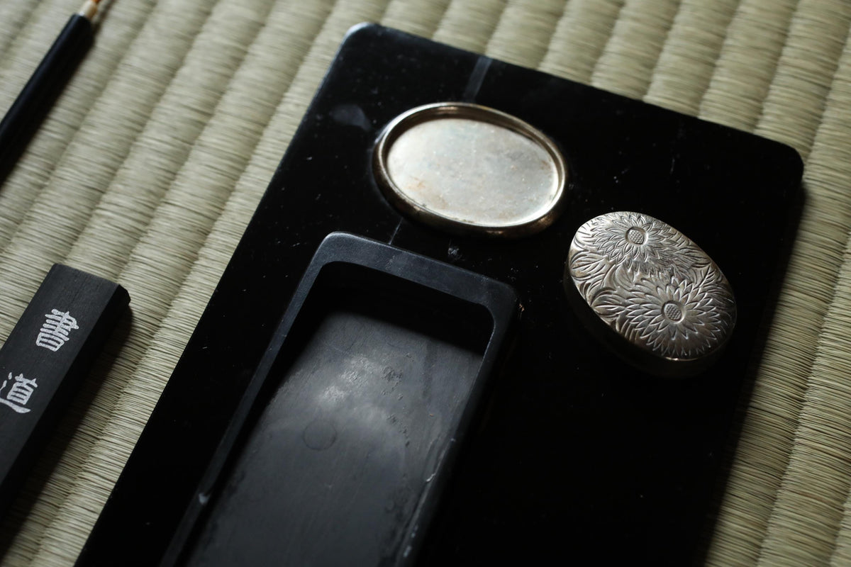 a black object with two metal components, one of which appears to be a small metal flower. The object is placed on a woven mat, and there is a small black object with Japanese characters next to it.