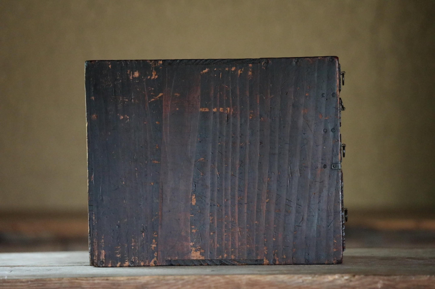 A weathered, dark wooden box sits on a wooden surface, its surface showing signs of age and use.