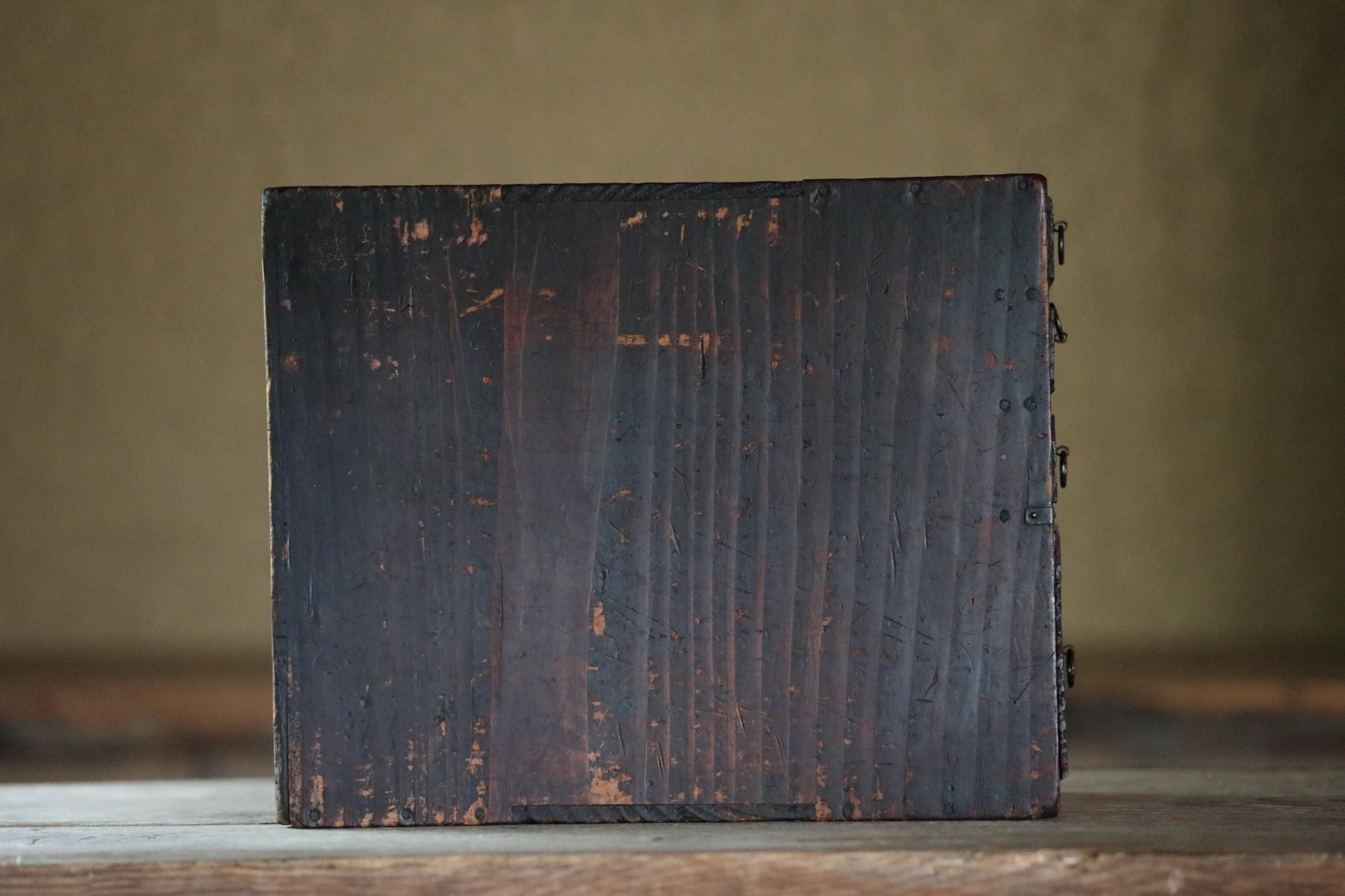 A weathered, dark wooden box sits on a wooden surface, its surface showing signs of age and use.