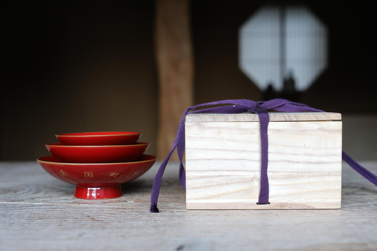 a wooden box with a purple ribbon tied around it, placed on a wooden surface next to a stack of red bowls.