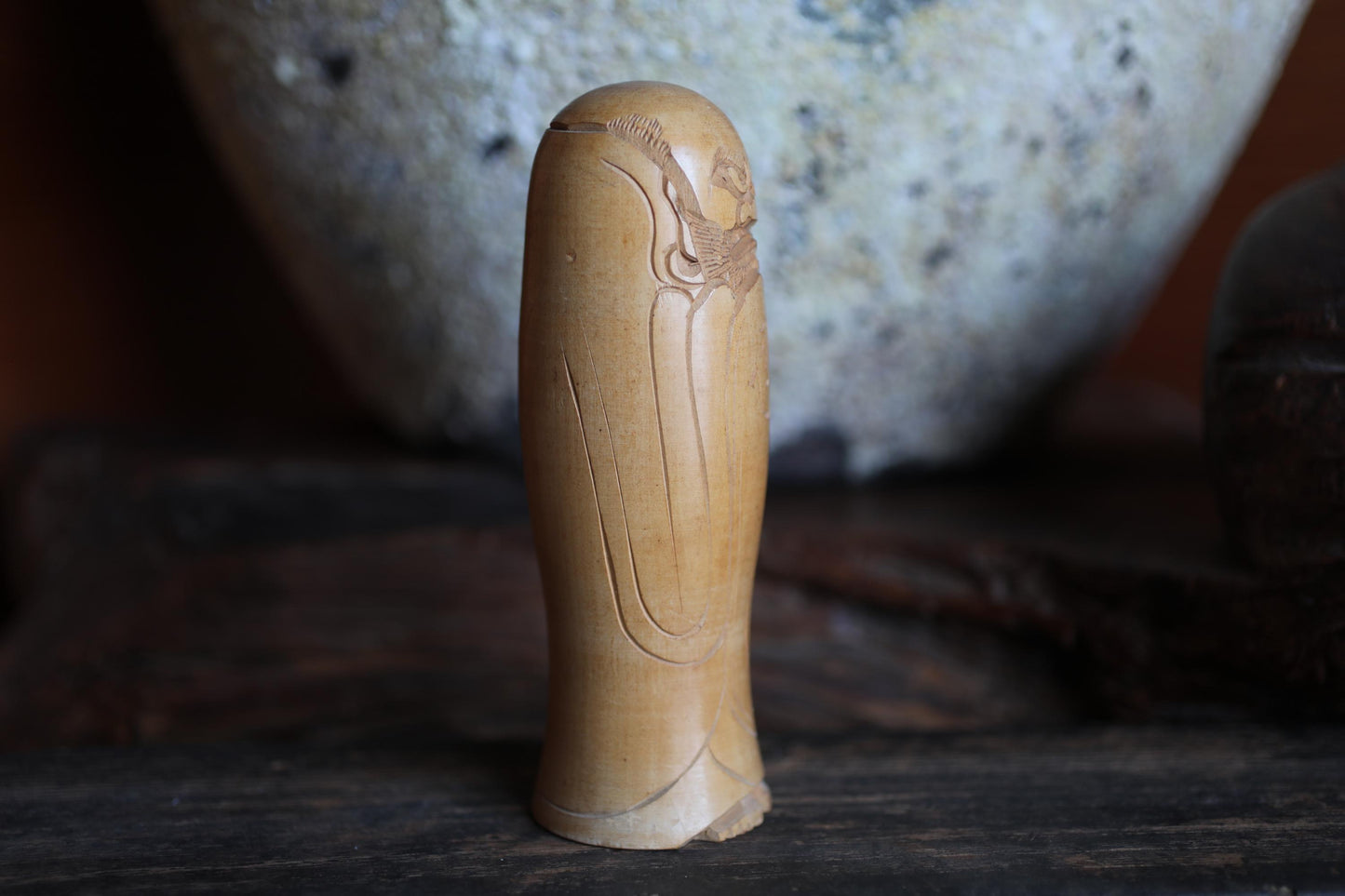 A wooden figurine with an intricate design is displayed on a wooden surface, with a large ceramic bowl visible in the background.