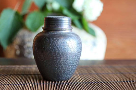 A small, round metal jar with a lid sits on a bamboo mat, with a blurred background featuring green leaves and a white flower.