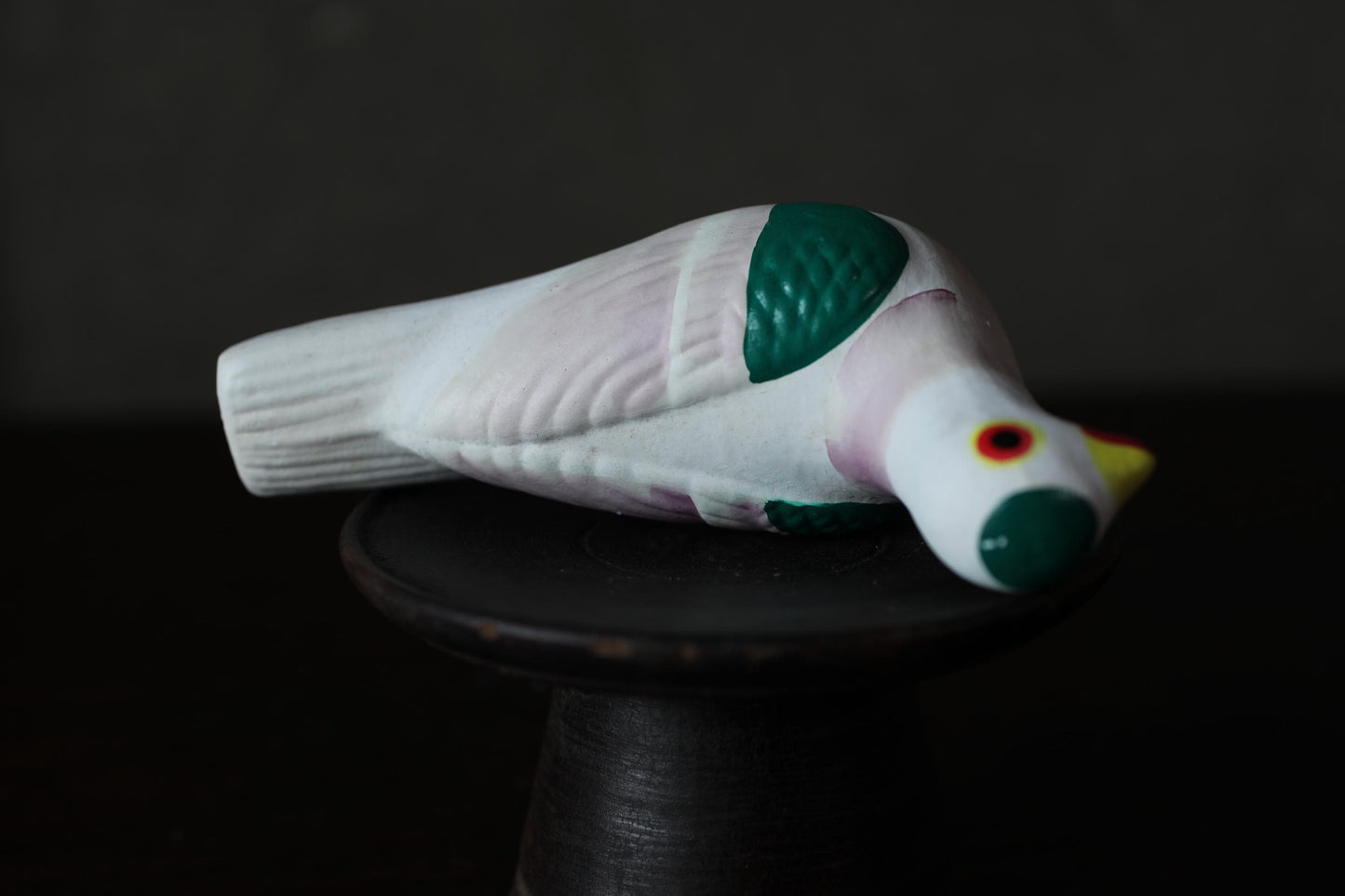 A ceramic bird figurine with a white body, green accents, and a red eye is sitting on a black stand against a dark background.