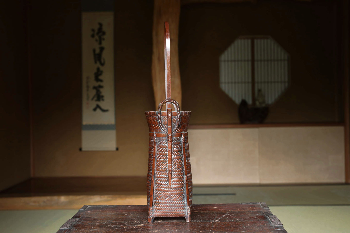 A wooden basket with a handle, placed on a wooden surface, with a traditional Japanese-style room in the background.