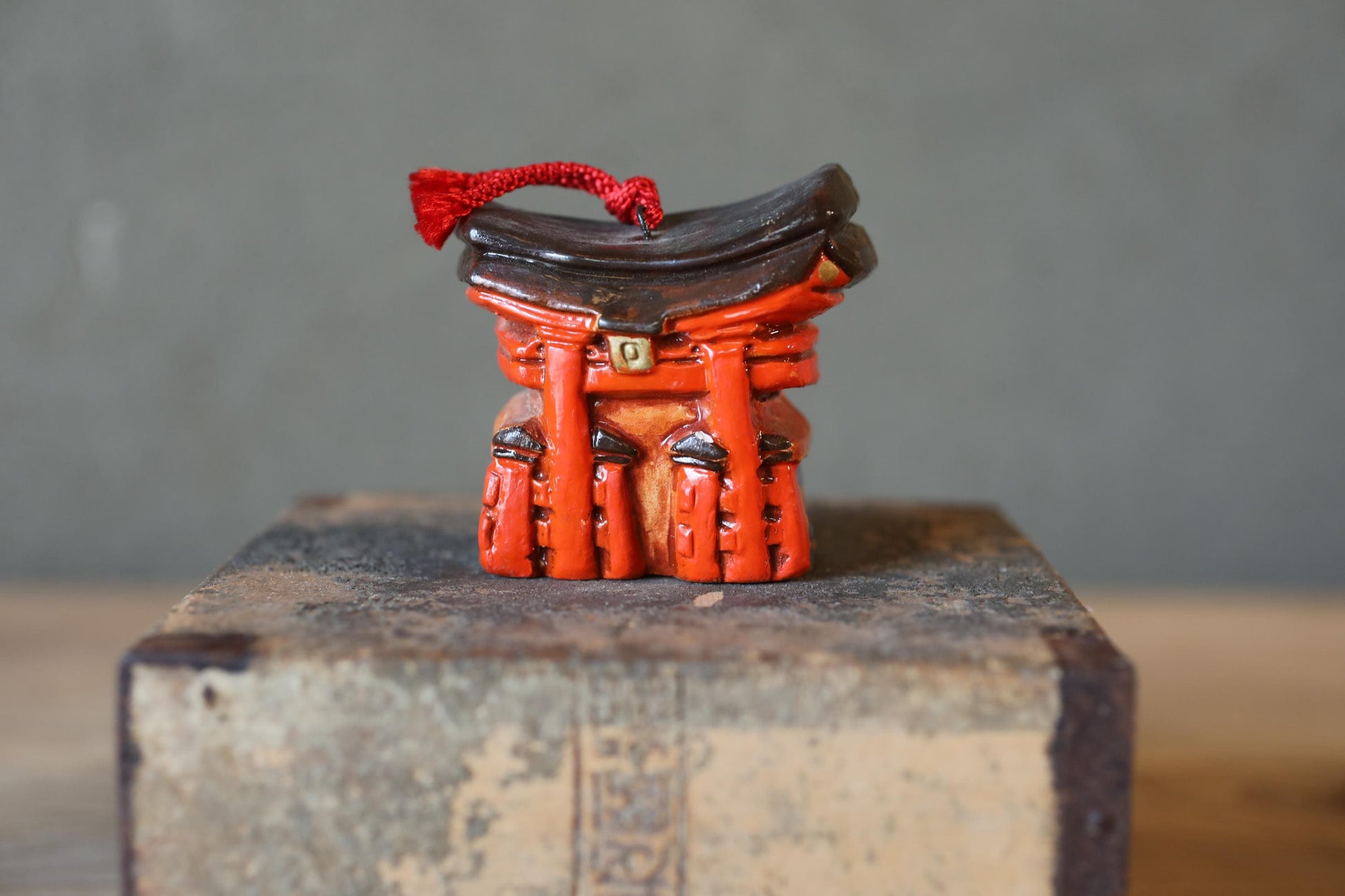 A small, red figurine of a shrine or shrine-like structure sits on a wooden surface against a gray background.