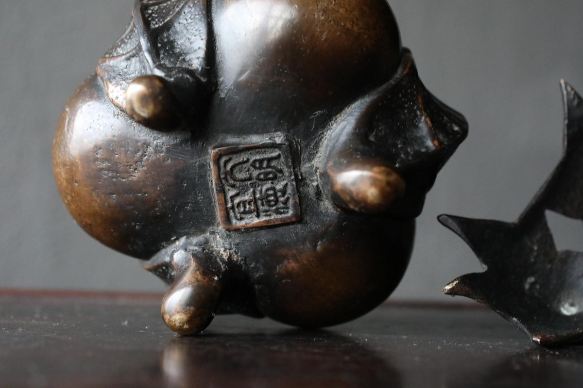 A close-up image of a bronze sculpture with a bird-like design, featuring a square emblem or stamp on the back.