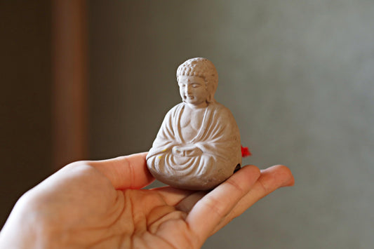 A small white Buddha statue is being held in a person's hand against a blurred background.