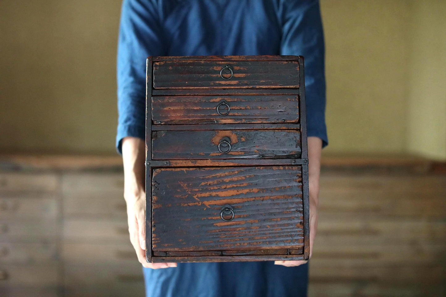 A person wearing a blue shirt is holding a wooden chest with four drawers in front of them.
