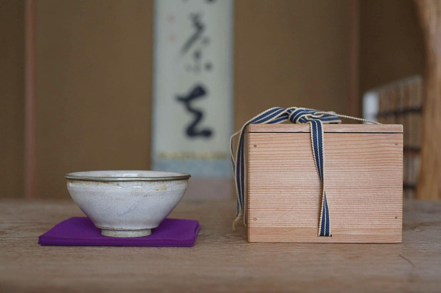 A white ceramic bowl and a wooden box with a blue ribbon tied around it are placed on a wooden table, with a blurred background that includes a wall with Japanese characters.
