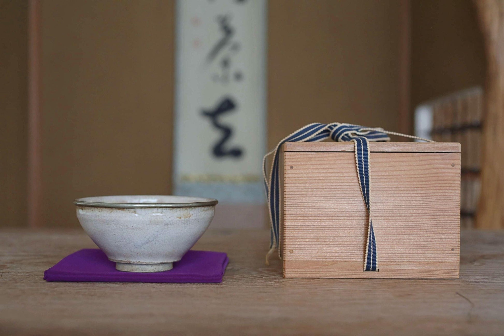 A white ceramic bowl and a wooden box with a blue ribbon tied around it are placed on a wooden table, with a blurred background that includes a wall with Japanese characters.