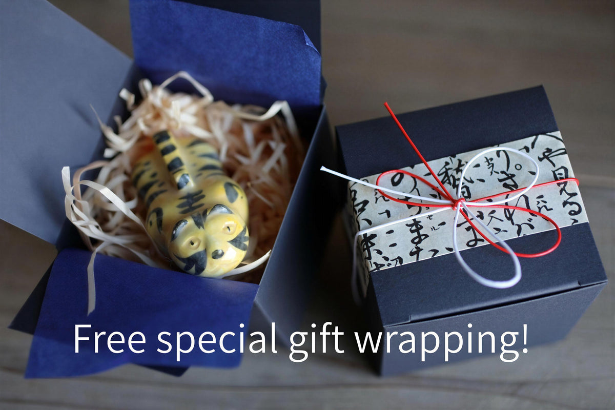 A small toy tiger figurine is nestled in a blue box filled with straw, accompanied by a black gift box with Japanese characters and a red ribbon tied around it.