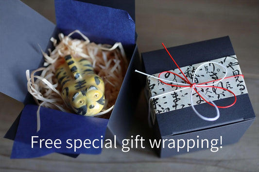 A small toy tiger figurine is nestled in a blue box filled with straw, accompanied by a black gift box with Japanese characters and a red ribbon tied around it.