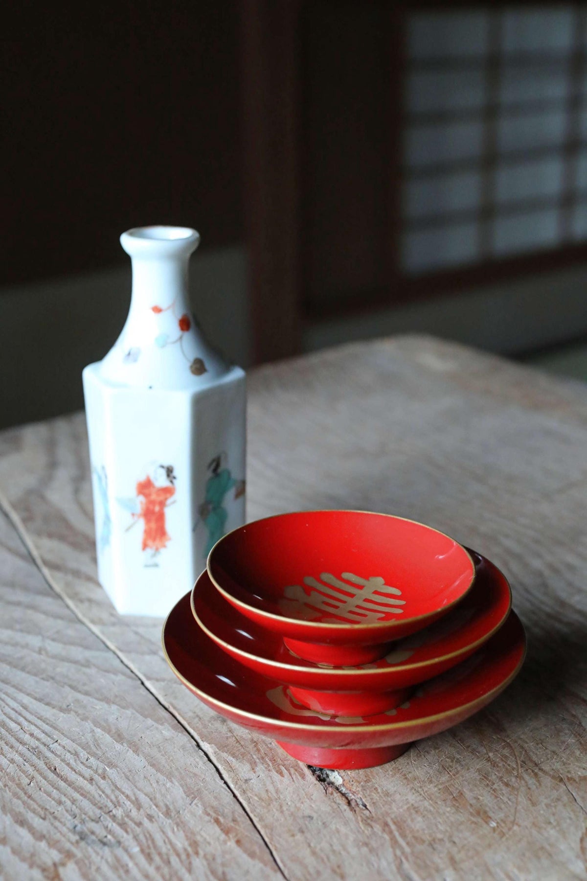 A white vase with a floral design and a stack of red bowls with gold trim are placed on a wooden table.