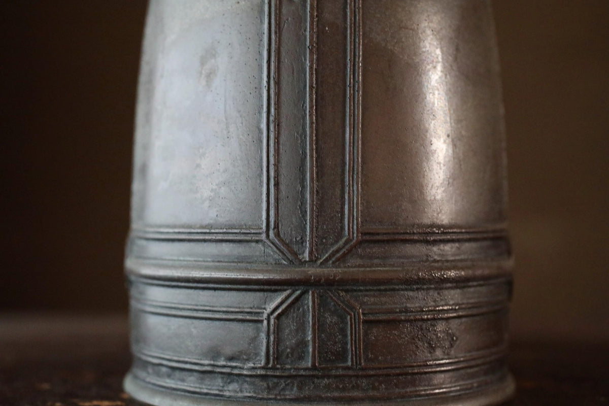A close-up view of a metal object, possibly a vase or container, with a textured surface and a geometric pattern.