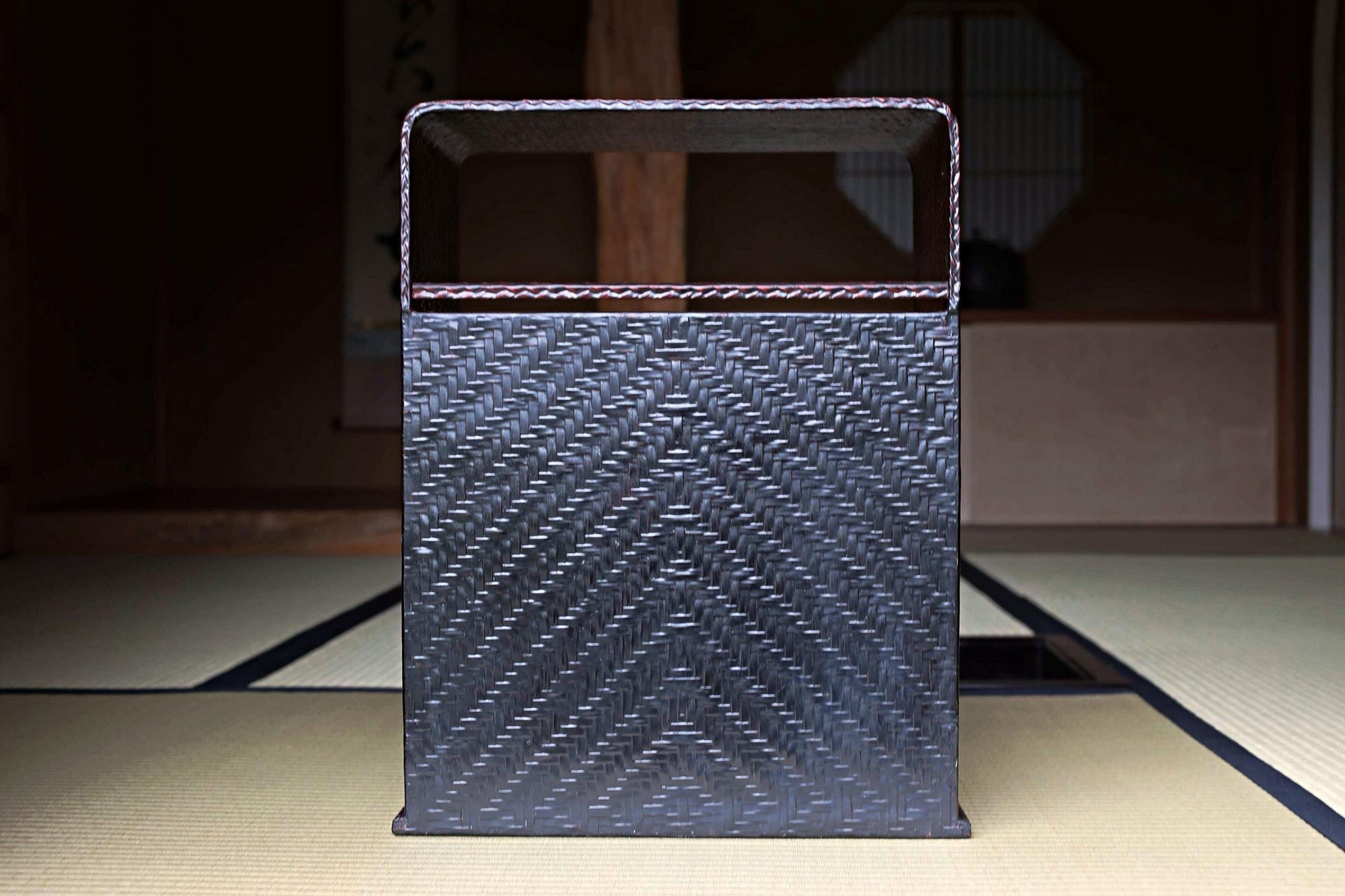 A black woven basket with a curved handle, placed on a tatami mat in a dimly lit room.